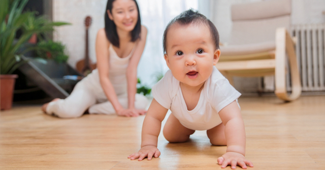 A photo of a crawling Asian American baby crawling toward the camera with their mother sitting on the floor in the background