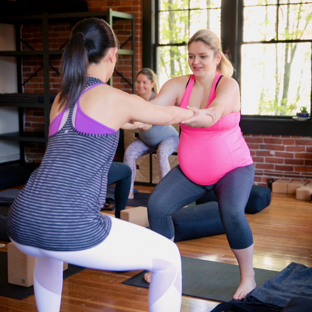 A photo of two pregnant women in the foreground (and two in the background) practicing a Patrner Squat in a prenatal yoga class at MamaSpace Yoga