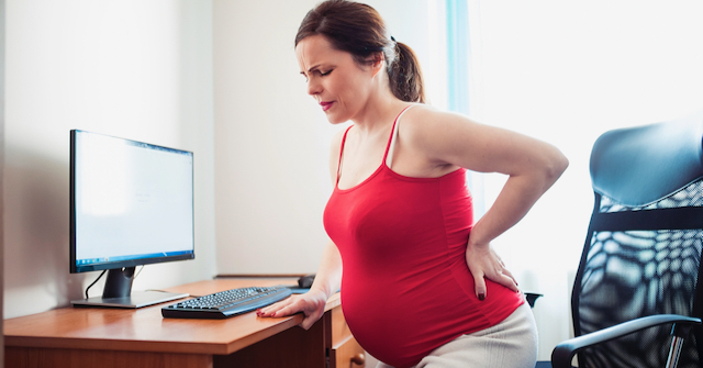 A photoi of a pregnant woman sitting in a chair at a desk holding her low back with a painful expression on her face