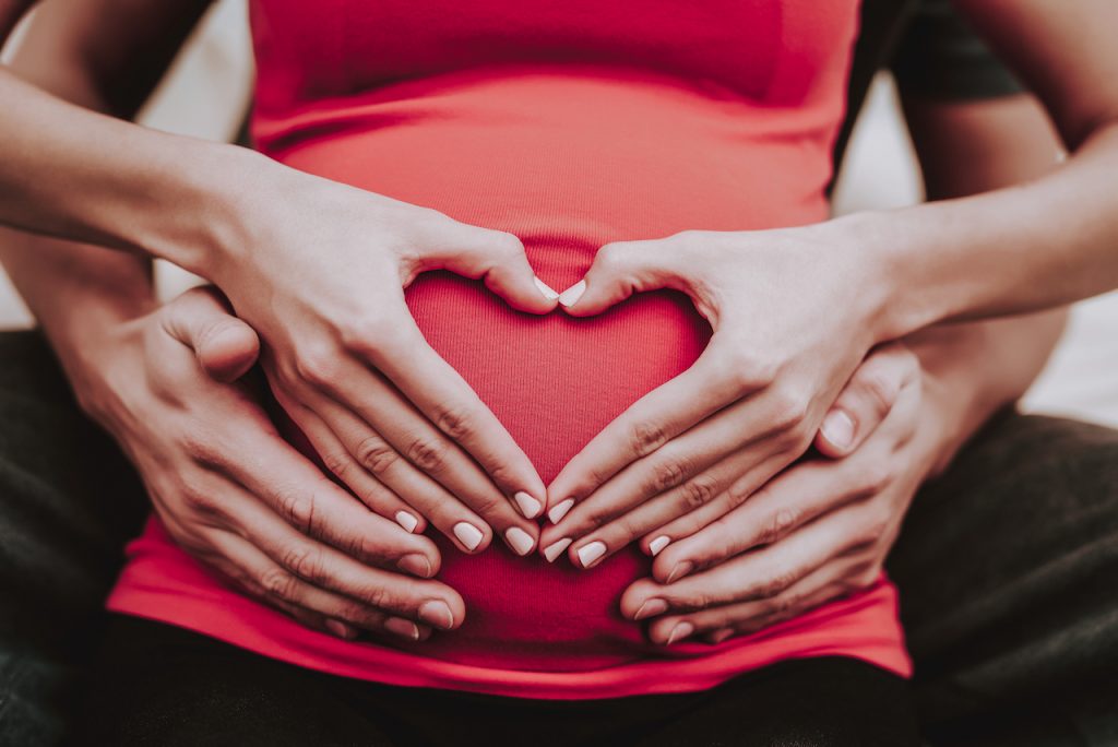 Two sets of hands make a heart shape on a pregnant belly
